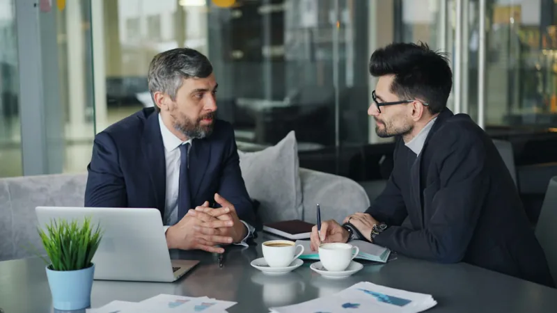 Twee mannen in pak in gesprek aan een tafel met een laptop